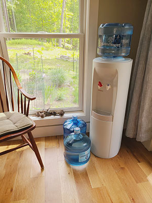A water cooler with a five-gallon bottle inserted in the top and two reserve bottles sitting on the floor next to it, with a window in the background looking out on a forest.